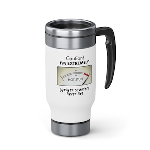 May include: White travel mug with a black handle. The mug has a graphic of a gauge with a red needle pointing to the word "HOT STUFF" and the text "Caution! I'M EXTREMELY (geiger counters never lie)"