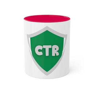 May include: White ceramic mug with a red rim. The mug features a green shield with a white outline and the text "CTR" in white.