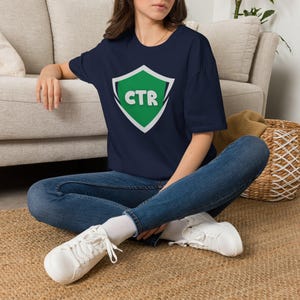 May include: A navy blue short-sleeve t-shirt featuring a central graphic of a green shield with a white border. The letters CTR are printed in white, bold, sans-serif font inside the shield. The shirt has a crew neckline and a relaxed fit.