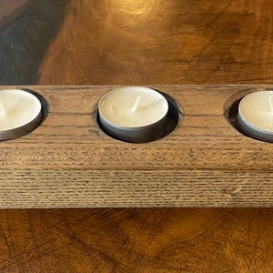 May include: A wooden candle holder with three circular recesses, each holding a small, round, white candle. The holder is rectangular and made of light brown wood, with a natural grain. The candles are in silver metal containers.
