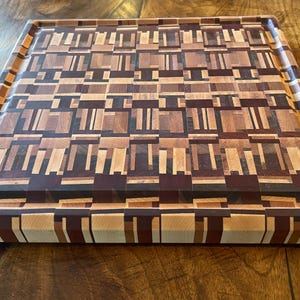 May include: A rectangular wooden chopping board featuring an intricate end-grain mosaic pattern. The surface displays a geometric arrangement of light, medium, and dark brown wood blocks, finished with a recessed juice groove along the perimeter.