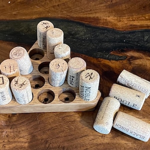 May include: A wooden wine cork holder with a triangular design, holding various wine corks. The corks display printed text and logos, including "Noble Vines" and "Black Birch". The holder and corks are on a wooden surface.