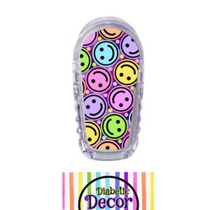 May include: A clear plastic case with a colorful sticker featuring a pattern of smiling faces in various colors on a purple background. The sticker is for a medical device. The text "Diabetic Decor by Danica" is on a rainbow striped background.