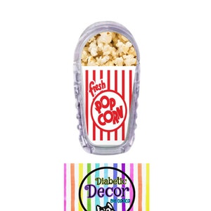 May include: A clear plastic insulin pump cover with a red and white striped design featuring the words "fresh pop corn". The cover has a picture of popcorn inside the container. The image also includes a logo for "Diabetic Decor by Damica" with a rainbow striped background.