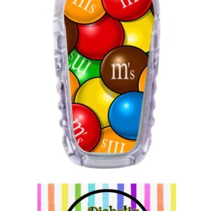 May include: A clear plastic insulin pump cover with a colorful design of M&M's candies. The design features red, yellow, blue, green, brown, and orange M&M's with the "m's" logo on each candy. The cover is for a diabetic pump and is sold by Diabetic Decor by Danica.