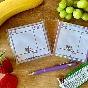 May include: Two colorful notepads with a cartoon husky dog on each. The notepads are labeled "Food", "Carbs", and "Insulin". The notepads are on a wooden surface with a banana, grapes, strawberries, a pen, and a granola bar.
