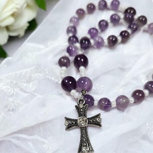 Puede incluir: Un collar de cuentas de amatista púrpura y blancas con un colgante de cruz de plata. La cruz tiene un strass transparente en el centro.