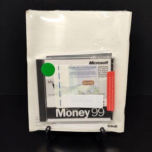 May include: Sealed Microsoft Money 99 software package. The package includes a CD-ROM, a certificate of authenticity, and a red warning label. The product is in a clear plastic wrap and is displayed on a black stand.