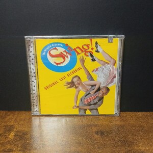 Puede incluir: Una caja de CD con el musical "Swing!". La portada tiene un fondo amarillo con el título en rojo y blanco, y un círculo azul. La imagen muestra bailarines en poses de swing. El texto dice "The Original Broadway" y "Original Cast Recording."