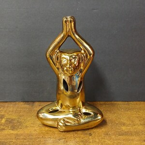 May include: A golden monkey figurine in a seated yoga pose. The figurine has its hands clasped above its head. The monkey's face is detailed with eyes, a nose, and a mouth. The figurine is on a wooden surface.