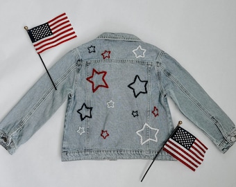 May include: Light blue denim jacket with embroidered stars in red, white, and navy blue. Two American flags are placed on either side of the jacket. The jacket is a classic style with a button-up front and collar.
