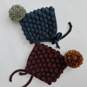 May include: Two crocheted baby hats with pom poms. One hat is dark blue with a green pom pom and the other is burgundy with a brown pom pom.
