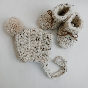 May include: A white crocheted baby hat with a pom-pom on top and matching booties with brown ribbon ties.