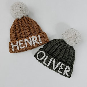 May include: Two crocheted hats with pom-poms. One hat is brown with the name "HENRI" embroidered on it. The other hat is green with the name "OLIVER" embroidered on it.