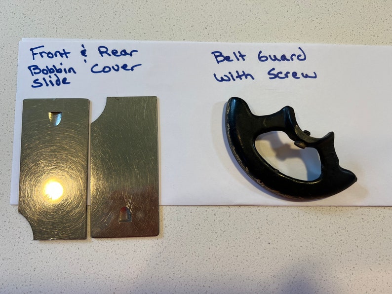 May include: Two silver metal plates with a small hole in each, labeled "Front & Rear Bobbin Cover Slide". A black plastic belt guard with a screw, labeled "Belt Guard with Screw".
