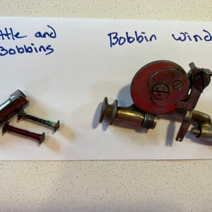 May include: A vintage sewing machine bobbin winder with a red metal spool and a silver metal base.  There is also a silver metal shuttle and two bobbins with thread.