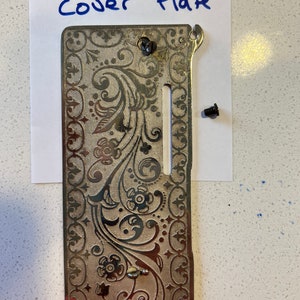 May include: A silver sewing machine front cover plate with a decorative floral design. The plate has a slot for the needle and a small hole for the bobbin. The text "Front Cover Plate" is written on a piece of paper behind the plate.