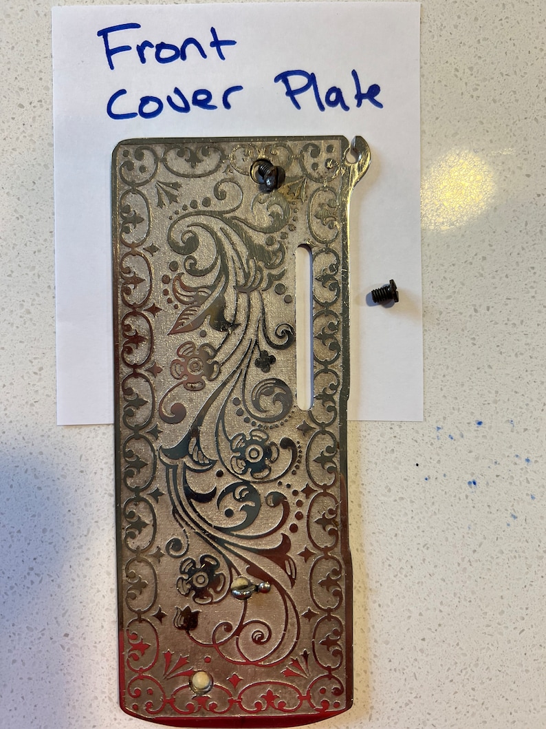 May include: A silver sewing machine front cover plate with a decorative floral design. The plate has a slot for the needle and a small hole for the bobbin. The text "Front Cover Plate" is written on a piece of paper behind the plate.