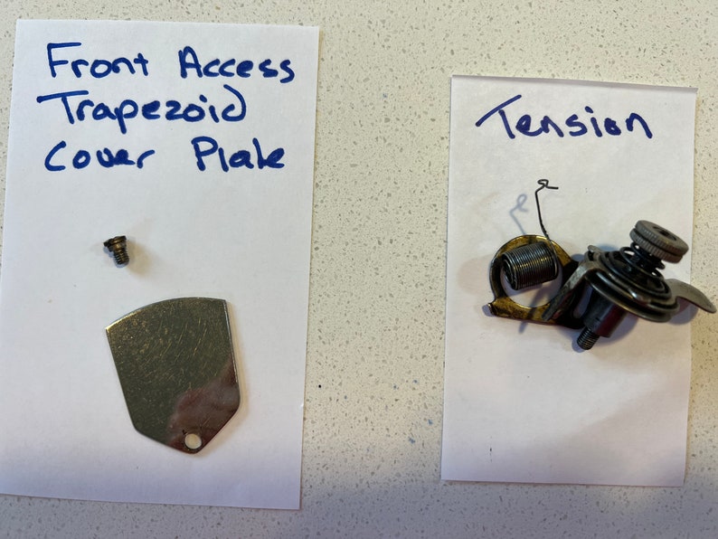 May include: Two pieces of a sewing machine: a metal cover plate with a screw and a tension assembly with a spring.