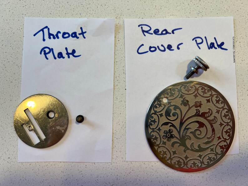 May include: Two sewing machine parts, a silver throat plate with a slot and a silver rear cover plate with a floral design. The rear cover plate has a small silver screw.