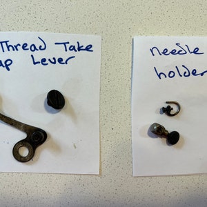 May include: Two pieces of paper with handwritten labels and sewing machine parts. The first piece of paper says "Thread Take up Lever" and has a metal lever and a black screw. The second piece of paper says "Needle holder" and has a metal needle holder, a small metal hook, and a screw.
