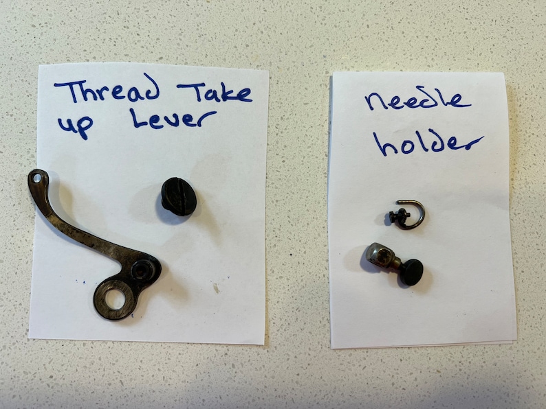 May include: Two pieces of paper with handwritten labels and sewing machine parts. The first piece of paper says "Thread Take up Lever" and has a metal lever and a black screw. The second piece of paper says "Needle holder" and has a metal needle holder, a small metal hook, and a screw.