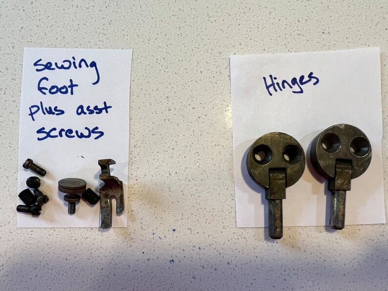 May include: Two silver-toned hinges with a round top and a metal pin. The hinges are on a white piece of paper with the word "Hinges" written in blue ink.  A separate piece of paper with the words "Sewing foot plus asst screws" written in blue ink.  There are several small screws and a sewing foot on the paper.