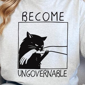 Könnte beinhalten: Hellgraues T-Shirt mit einer schwarzen Grafik einer Katze, die in eine Hand beißt, umrahmt von einem Rechteck. Die Worte "BECOME" und "UNGOVERNABLE" sind oberhalb und unterhalb der Grafik aufgedruckt.