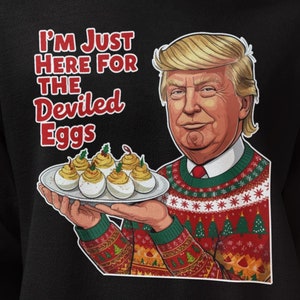 May include: Black hooded sweatshirt featuring a cartoon image of a man holding a plate of deviled eggs. The man is wearing a Christmas sweater. Text on the sweatshirt reads "I'M JUST HERE FOR THE Deviled Eggs."