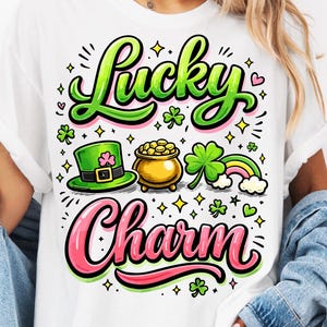 May include: White t-shirt with a colorful "Lucky Charm" graphic. The design includes a green leprechaun hat, a pot of gold, a four-leaf clover, and a rainbow. The text is in green and pink, with a playful font, perfect for St. Patrick's Day.