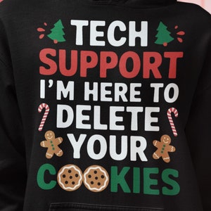 以下が含まれることがあります： 黒いフード付きスウェットシャツ。赤、白、緑で「TECH SUPPORT I'M HERE TO DELETE YOUR COOKIES」の文字。デザインにはクリスマスツリー、ジンジャーブレッドマン、キャンディケーン、クッキーが含まれています。