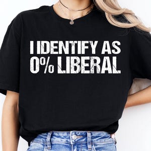 May include: Black t-shirt with the white text "I IDENTIFY AS 0% LIBERAL." The shirt is worn with blue jeans. A plant is visible in the background.