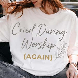 Puede incluir: Camiseta blanca con el texto "Cried During Worship (AGAIN)" en escritura gris y dorada. La camiseta presenta un diseño botánico. La persona de la imagen lleva una gorra marrón y gafas de sol.