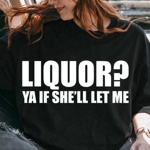 May include: Black t-shirt with the text "LIQUOR? YA IF SHE'LL LET ME" in white. The person in the image is wearing a brown hat and sunglasses. The person is sitting in a car.