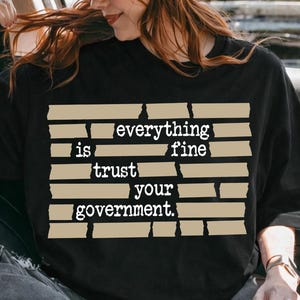 Può includere: T-shirt nero con la scritta "everything is fine trust your government" in un design beige a forma di nastro adesivo. La maglietta è indossata da una persona in auto, con un cappello marrone e occhiali da sole.