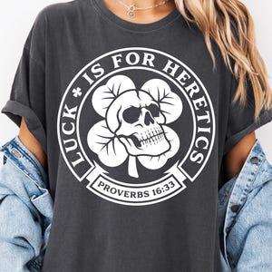 Peut inclure: T-shirt gris foncé avec un motif blanc. Le motif est un crâne à l'intérieur d'un trèfle à quatre feuilles, entouré des mots "LUCK IS FOR HERETICS" et "PROVERBS 16:33". Le t-shirt a des manches courtes et une coupe décontractée.