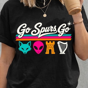 K&ouml;nnte beinhalten: Schwarzes T-Shirt mit dem Text "Go Spurs Go" in Wei&szlig;, mit einer teal-farbenen Fuchs-, pinkfarbenen Alien-, orangefarbenen Burg- und wei&szlig;en Harfengrafik. Das Shirt hat einen Rundhalsausschnitt und kurze &Auml;rmel.