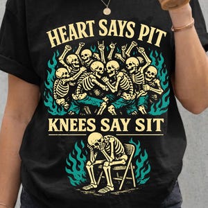 Può includere: T-shirt nero con la scritta "HEART SAYS PIT KNEES SAY SIT" e una grafica di scheletri. La grafica include scheletri che ballano e uno scheletro seduto su una sedia, con fiamme turchesi.