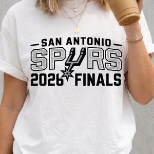 Puede incluir: Una camiseta blanca de manga corta con el texto negro SAN ANTONIO SPURS 2026 FINALS y el logotipo del equipo integrado en la palabra SPURS. La camiseta se combina con pantalones cortos de mezclilla gris oscuro y un collar dorado.