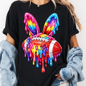 May include: Black t-shirt featuring a colorful graphic of a football designed as a bunny with rainbow-colored ears and dripping paint. The shirt is a casual, oversized style.