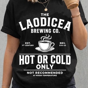 May include: A black short-sleeved t-shirt featuring white graphic text that reads THE LAODICEA BREWING CO. EST. 1ST CENTURY REV. 3:14-22 HOT OR COLD ONLY NOT RECOMMENDED AT ROOM TEMPERATURE, with a central illustration of a steaming coffee cup.