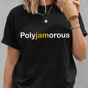 Peut inclure: T-shirt noir avec le mot "Polyamorous" en lettres blanches et jaunes. Le t-shirt est porté avec un short en jean noir. La personne tient une tasse en plastique transparente avec une boisson brune et une paille.