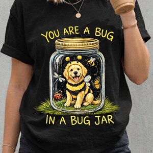 Op de afbeelding: Een zwart t-shirt met korte mouwen en een afbeelding van een gouden puppy in een bijenkostuum in een glazen pot. De tekst "YOU ARE A BUG IN A BUG JAR" staat in het geel boven en onder de pot, die ook een lieveheersbeestje en vlinder bevat.