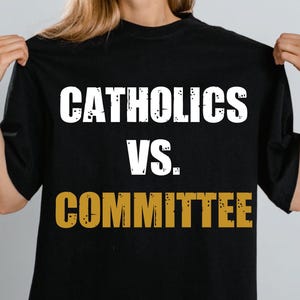 Op de afbeelding: Zwart T-shirt met de tekst "CATHOLICS VS. COMMITTEE" in witte en gouden letters. Het shirt wordt door een persoon omhoog gehouden.