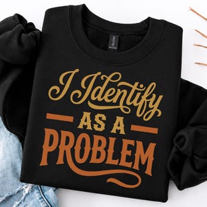 May include: A black crewneck sweatshirt featuring the text "I IDENTIFY AS A PROBLEM" printed in a stylised, bold orange and gold font. The garment has long sleeves and a ribbed neckline, presented alongside a pair of light blue distressed denim jeans.