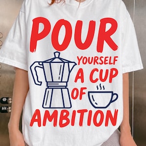 Puede incluir: Una camiseta blanca de manga corta con un estampado gráfico en rojo y azul oscuro. El diseño incluye una cafetera italiana azul oscuro y una taza de café humeante, con el texto rojo: POUR YOURSELF A CUP OF AMBITION.