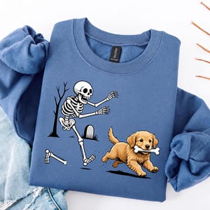 May include: A blue crewneck sweatshirt featuring a graphic print of a white skeleton chasing a golden puppy that is running with a bone in its mouth. The skeleton is depicted near a tombstone and a bare tree, with one detached leg bone nearby.