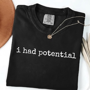 Könnte beinhalten: Schwarzes T-Shirt mit Rundhalsausschnitt und dem weißen Text "i had potential". Eine goldfarbene Halskette mit rundem Anhänger liegt auf dem Shirt. Das Shirt hat ein Comfort Colors-Etikett. Ein brauner Hut und dekorative Federn sind im Hintergrund.