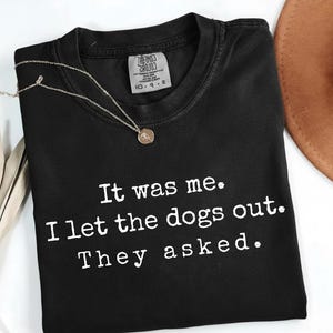 May include: Black crew neck t-shirt with the text "It was me. I let the dogs out. They asked." in white. A silver necklace with a pendant is on the shirt. A brown hat is in the background.