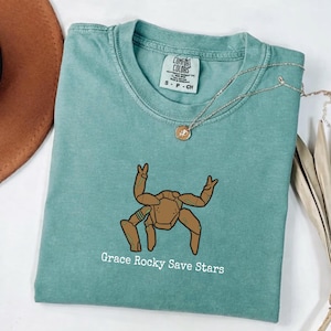 May include: A sage green short-sleeve t-shirt featuring a brown, multi-legged creature graphic with the text "Grace Rocky Save Stars" printed in white below it. The shirt has a Comfort Colors brand label at the inner neckline and a crew neck collar.